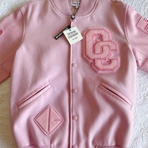 Puede incluir: Una chaqueta de béisbol rosa con un gran parche rosa "OC" en la parte delantera. La chaqueta tiene un cuello, puños y cintura de canalé. La chaqueta tiene dos bolsillos en la parte delantera.
