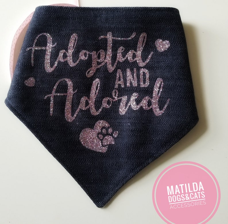 May include: A black denim dog bandana with pink glitter lettering that says "Adopted and Adored" with a pink glitter heart and paw print.
