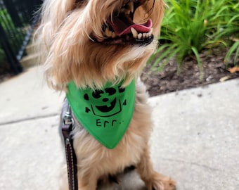 May include: A small, light brown dog wearing a green bandana with a black and white graphic and the text "Err."