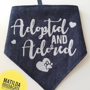 May include: A dark blue denim dog bandana with silver glitter lettering that says "Adopted and Adored" with a silver glitter paw print heart.