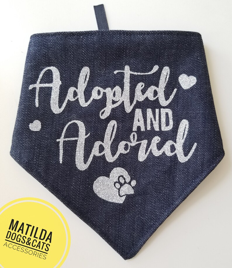 May include: A dark blue denim dog bandana with silver glitter lettering that says "Adopted and Adored" with a silver glitter paw print heart.