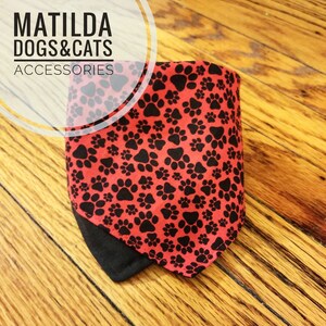 May include: Red bandana with a black paw print pattern. The bandana has a black border and is folded in half. The bandana is on a wooden surface. The text "MATILDA DOGS & CATS ACCESSORIES" is visible in the top left corner of the image.