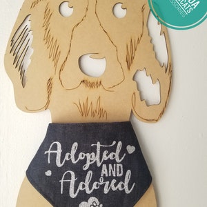 May include: A blue denim dog bandana with silver glitter text that reads "Adopted and Adored" with a silver glitter paw print heart. The bandana is on a wooden dog cutout.