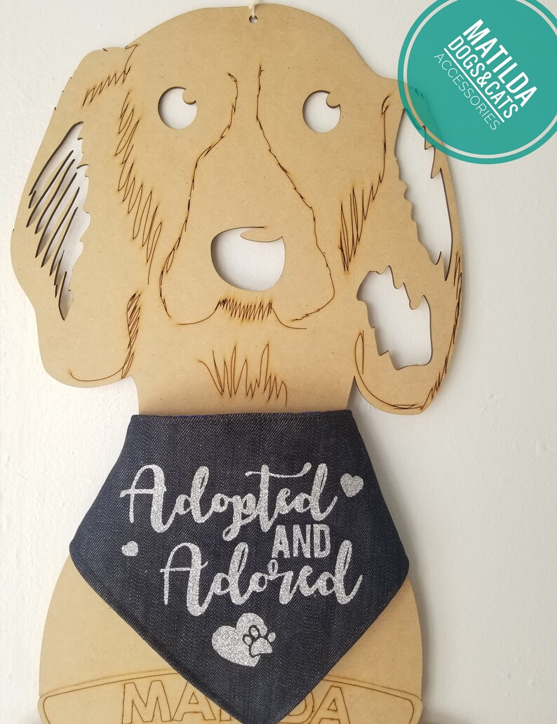 May include: A blue denim dog bandana with silver glitter text that reads "Adopted and Adored" with a silver glitter paw print heart. The bandana is on a wooden dog cutout.