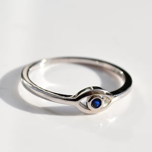 May include: A silver ring featuring a blue eye charm. The ring is a simple band with a small eye charm that has a blue stone in the centre.