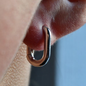 Può includere: Orecchini a cerchio in argento con un designo semplice e arrotondato. Gli orecchini sono mostrati nell'orecchio di una persona.
