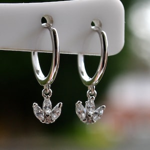 May include: A pair of silver hoop earrings with a dangling flower-shaped charm featuring clear cubic zirconia stones.