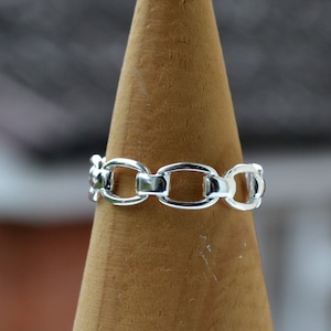 May include: A silver chain ring with oval links. The ring is displayed on a wooden cone.