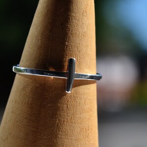May include: A silver ring with a simple cross design. The ring is displayed on a wooden ring holder.