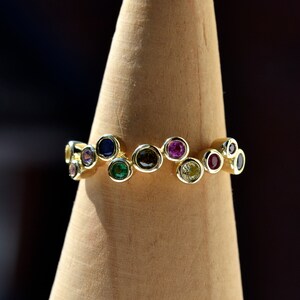 May include: A gold ring featuring a row of colourful gemstones. The gemstones are set in a wavy pattern and include green, blue, purple, red, and yellow.