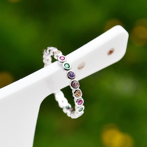 Peut inclure: Une bague en argent avec une bande de pierres précieuses colorées. Les pierres précieuses sont serties en cercle autour de la bande et forment un arc-en-ciel.