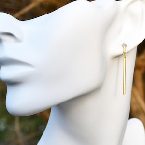 May include: A single gold-coloured bar earring is shown on a white mannequin head. The earring hangs vertically, suspended from a small loop at the top. The background is blurred, with hints of green and brown.