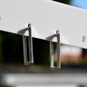 May include: Two silver rectangular hoop earrings suspended from a white display. The earrings feature a minimalist, open design with a shiny finish. The background is blurred, implying an outdoor setting.
