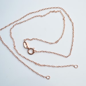 May include: A delicate rose gold chain necklace with a lobster clasp and two open jump rings. The chain is made of small, interconnected links and is displayed against a white background.