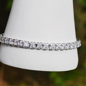 May include: A silver-toned tennis bracelet featuring a continuous row of clear, rectangular-cut stones. The bracelet is displayed wrapped around a white conical stand, showcasing the uniform arrangement and reflective quality of the faceted stones.