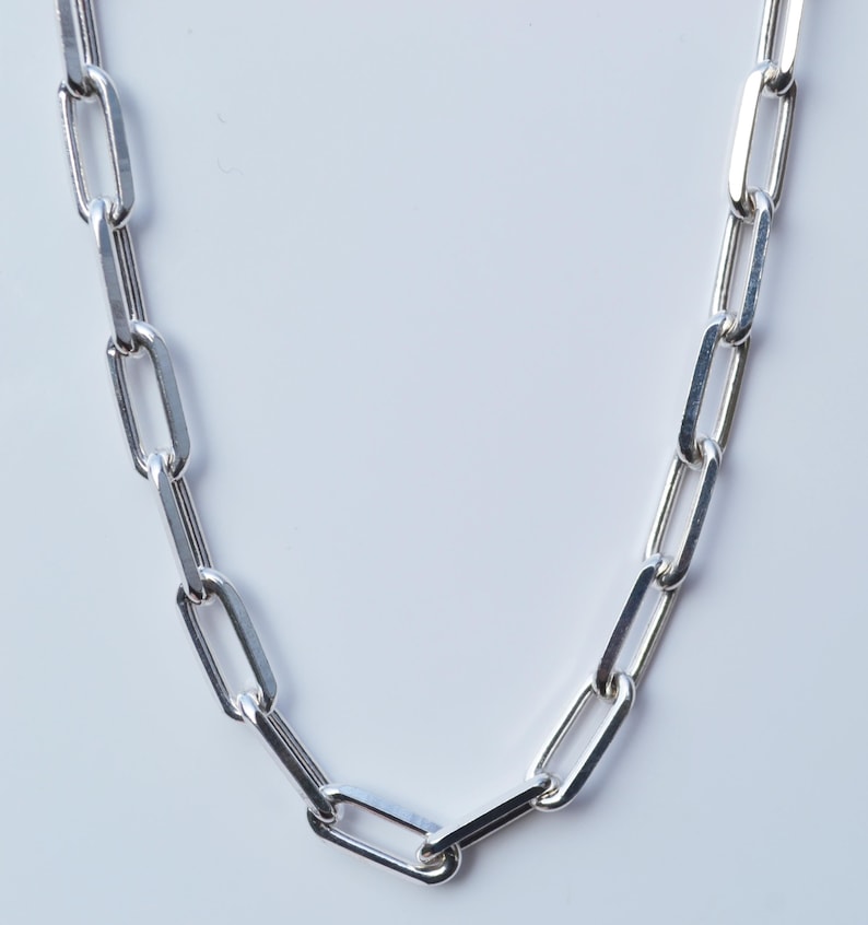 May include: A silver chain necklace with a link style that resembles a paperclip. The chain is made of polished metal and has a smooth, shiny finish.