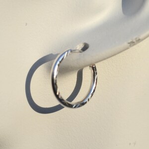 May include: A silver hoop earring with a textured, diamond-cut design. The earring is shown in a close-up view, highlighting the intricate pattern and the shiny metal. The earring is attached to an ear.