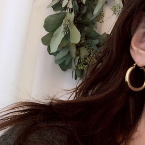 May include: Gold hoop earrings with a thick, rounded design. The earrings are worn by a person with long brown hair.