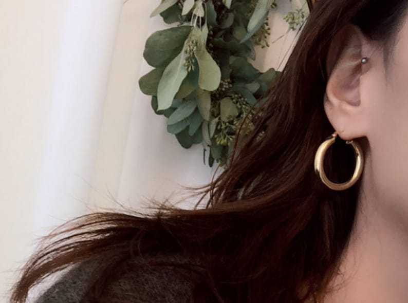 May include: Gold hoop earrings with a thick, rounded design. The earrings are worn by a person with long brown hair.