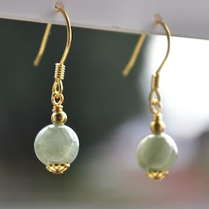 K&ouml;nnte beinhalten: Ein Paar goldene Ohrringe mit gr&uuml;nen Edelsteinperlen. Die Ohrringe haben ein schlichtes Design mit einer einzelnen Perle an jedem Ohrring.