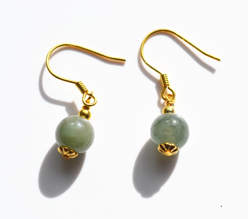 K&ouml;nnte beinhalten: Ein Paar goldene Ohrringe mit gr&uuml;nen Jade-Perlen. Die Ohrringe haben ein schlichtes Design mit einer kleinen goldenen Blumenverzierung an der Unterseite jeder Perle.