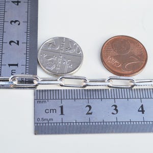 May include: A silver chain with a link size of 5 mm, measured against a ruler and two coins. The chain is made of silver and has a polished finish.