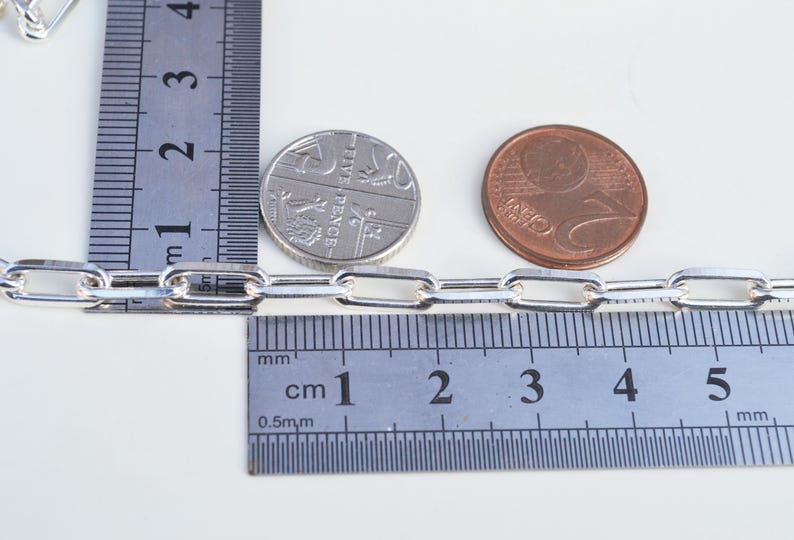 May include: A silver chain with a link size of 5 mm, measured against a ruler and two coins. The chain is made of silver and has a polished finish.