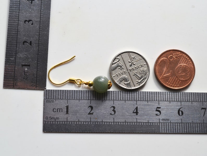 K&ouml;nnte beinhalten: Ein Paar gr&uuml;ne Jade-Ohrringe mit goldenen Haken. Die Ohrringe werden neben einem Lineal und zwei M&uuml;nzen gezeigt, einer britischen F&uuml;nf-Pence-M&uuml;nze und einer 20-Cent-Euro-M&uuml;nze, als Gr&ouml;&szlig;envergleich.