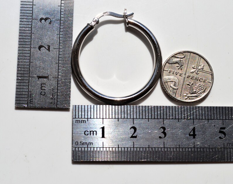 May include: Silver hoop earrings with a diameter of approximately 2.5 cm, shown next to a ruler and a British five pence coin.