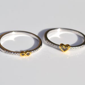 May include: Two silver rings with gold detailing. One ring has a gold infinity symbol, the other a gold heart. Both rings have a band of small, clear stones. The rings are set against a white background.