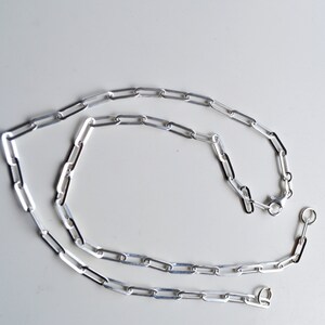 May include: A silver chain necklace with a lobster clasp and a jump ring. The chain is made of small, rectangular links.