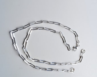 May include: A silver chain necklace with a lobster clasp and a jump ring. The chain is made of small, rectangular links.