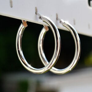 May include: A pair of silver hoop earrings hanging on a white display stand. The earrings are simple and classic, with a smooth, polished finish.