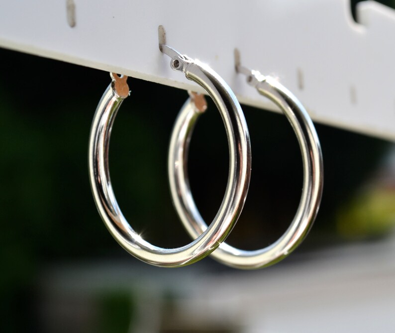 May include: A pair of silver hoop earrings hanging on a white display stand. The earrings are simple and classic, with a smooth, polished finish.