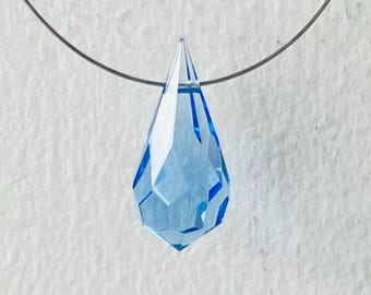 May include: A faceted, light blue crystal pendant necklace. The teardrop-shaped pendant is suspended from a thin, silver-colored chain. The crystal has multiple facets that catch the light, creating a sparkling effect. The background is a textured, off-white surface.