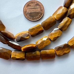 May include: Close-up of strands of tiger eye gemstone beads. The beads are rectangular with faceted surfaces, displaying a rich, golden-brown colour. A US one-cent coin is included for size reference. The beads are strung together, suggesting their use in jewellery making.