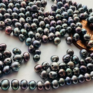 May include: A close-up of a pile of dark iridescent oval-shaped pearls with a UK penny for scale. The pearls are scattered on a white surface.
