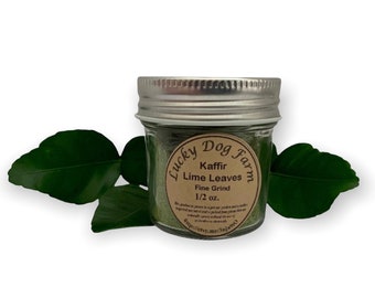 May include: A small glass jar with a silver lid filled with green kaffir lime leaves. The jar has a brown label that reads "Lucky Dog Farm" and "Kaffir Lime Leaves Fine Grind 1/2 oz."