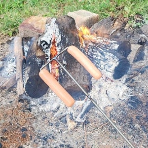 May include: Two hot dogs are being roasted over a campfire on a metal roasting stick. The hot dogs are partially cooked and have a golden brown color. The campfire is made of logs and is burning brightly.