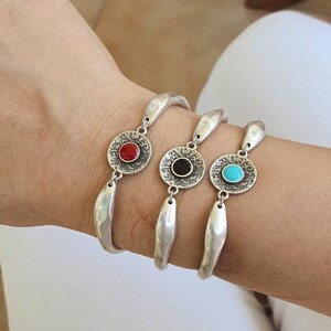 Può includere: Tre braccialetti d'argento con centri circolari decorativi. Ogni braccialetto presenta una pietra di colore diverso: rossa, nera e turchese. I braccialetti sono indossati al polso.