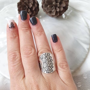 Può includere: Un anello color argento con un intricato design a mandala simmetrico. L'anello è largo e copre una parte significativa del dito. Lo sfondo include pigne e una superficie bianca.