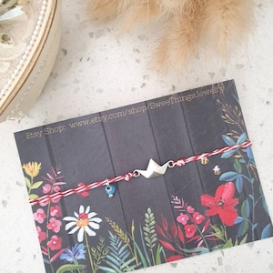 Puede incluir: Una pulsera hecha a mano con un dije de barco de papel, una cuenta azul de mal de ojo y una cuerda roja y blanca. La pulsera se exhibe en una tarjeta con ilustraciones florales y una URL de la tienda Etsy.