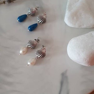 Puede incluir: Pendientes de plata con forma de concha, con cuentas azules en forma de lágrima y pendientes de perlas, presentados sobre un plato de mármol con ramitas de lavanda y piedras blancas. Joyas.