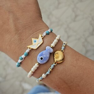 Op de afbeelding: Drie armbanden met nautische bedels. Eén armband heeft een goudkleurige papieren bootbedel, een andere heeft een blauwe visbedel en de derde heeft een gouden schelpenbedel. De armbanden zijn geregen met witte en turquoise kralen.