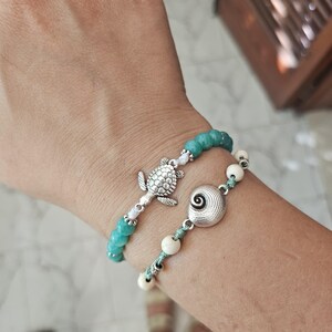 Peut inclure: Deux bracelets de perles. L'un présente des perles turquoise et un charm en argent en forme de tortue. L'autre a des perles blanches et un charm en argent en forme de spirale. Les deux bracelets sont sur un poignet.
