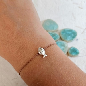 Könnte beinhalten: Armband mit einem silbernen Fischanhänger an einem braunen Band. Der Fischanhänger ist klein und detailliert. Das Armband wird am Handgelenk getragen, mit blau-grünen Steinen im Hintergrund.