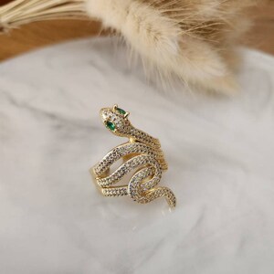 May include: A gold-colored snake ring with a coiled design. The ring is encrusted with small, clear crystals and features two green emerald-colored stones for eyes. The ring is set on a white marble surface.