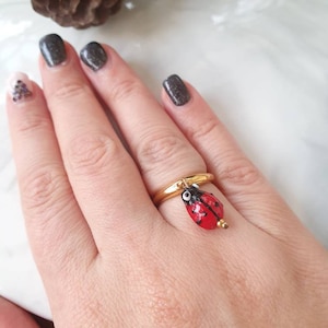 Könnte beinhalten: Ein goldfarbener Ring mit einem Anhänger in Form eines Marienkäfers. Der Marienkäfer ist rot mit schwarzen Punkten und einem schwarzen Kopf. Der Ring befindet sich an einem Finger, vor einem weißen und hellbraunen Hintergrund.