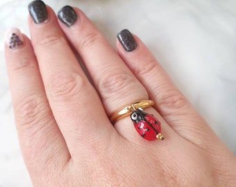 May include: A gold-colored ring with a dangling ladybug charm. The ladybug is red with black spots and a black head. The ring is on a person's finger, with a white and light brown background.