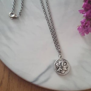 Könnte beinhalten: Eine silberfarbene Halskette mit einem Anhänger, der das Profil einer Person zeigt. Die Kette ist filigran und liegt auf einer weiß-grau marmorierten Oberfläche, mit lila Blumen im Hintergrund.